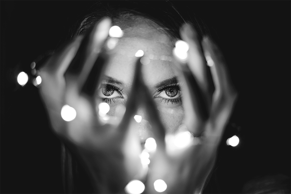 A woman looks at the camera through her hands, which are holding string lights